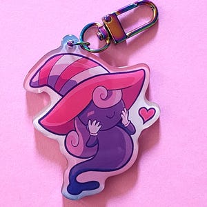 May include: A cartoon ghost keychain with a witch's hat. The ghost is purple, the hat is pink and striped, and there is a small pink heart. The clasp is rainbow-coloured.