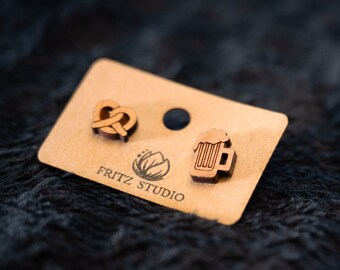 May include: A pair of wooden earrings with a pretzel and a beer mug design. The earrings are on a brown card with the text "Fritz Studio".