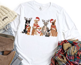 Puede incluir: Camiseta blanca de manga larga con un gráfico de un grupo de perros con cuernos de reno y gorros de Papá Noel. Los perros son de diferentes razas y tamaños.