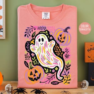 May include: A terracotta-coloured long-sleeved shirt with a whimsical ghost design, featuring colourful brushstrokes, pumpkins, and floral elements. The shirt has a crew neck and a "Comfort Colors" tag. The theme is Halloween.