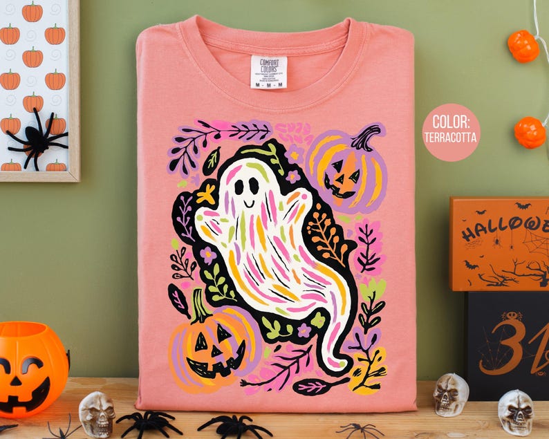 May include: A terracotta-coloured long-sleeved shirt with a whimsical ghost design, featuring colourful brushstrokes, pumpkins, and floral elements. The shirt has a crew neck and a "Comfort Colors" tag. The theme is Halloween.