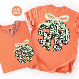 May include: Two melon-colored t-shirts with a green bow and a monogram design. The monogram is a circle with the letters S, M, and a stylized M, filled with a leopard print pattern and green shamrocks. One shirt has a silver chain necklace.