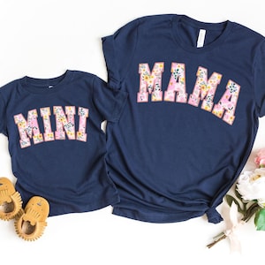 May include: Two navy blue t-shirts with floral print lettering. The larger shirt says "MAMA" and the smaller shirt says "MINI".