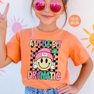 May include: Melon-colored t-shirt with the text "A Little Bit Dramatic" and a smiley face graphic. The graphic includes a pink hat, peace signs, and a checkered background. The shirt is worn by a person wearing pink sunglasses.