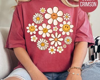 May include: A crimson red t-shirt with a graphic design of smiling daisies. The daisies are white with yellow centers and have a yellow smiley face in the center of each flower.