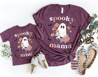 May include: Two heather maroon t-shirts with the words "spooky mama" and "spooky mini" in a retro font. Each shirt features a white ghost illustration surrounded by flowers and bats. The shirts are paired with matching shoes.