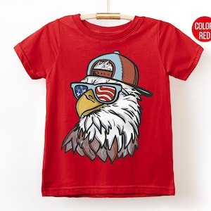 May include: Red t-shirt featuring a graphic of an eagle wearing sunglasses with an American flag design and a baseball cap. The eagle's head is detailed with white, brown, and yellow colors. The shirt is hanging on a wooden hanger.