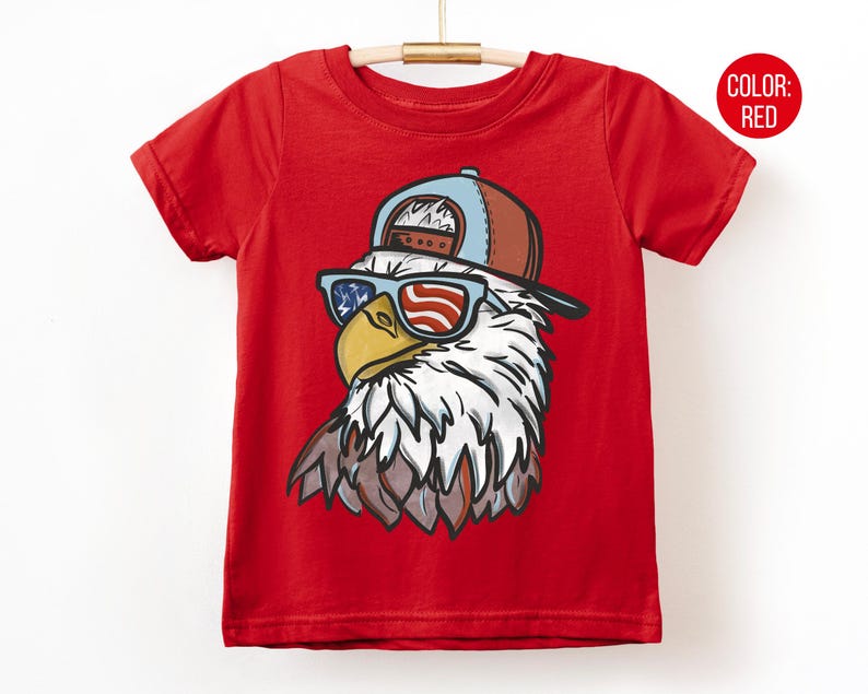 May include: Red t-shirt featuring a graphic of an eagle wearing sunglasses with an American flag design and a baseball cap. The eagle's head is detailed with white, brown, and yellow colors. The shirt is hanging on a wooden hanger.