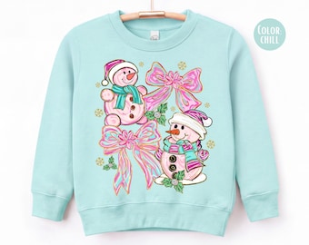 Peut inclure: Un sweat-shirt à col rond vert menthe clair orné d'un motif de deux bonhommes de neige roses portant des écharpes et des bonnets bleu sarcelle, avec de grands nœuds roses irisés, du houx vert et des flocons dorés. Le texte COLOR: CHILL est visible.
