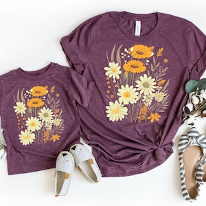 May include: Two short-sleeved t-shirts in a heathered plum color, each featuring a central graphic print of a wildflower bouquet with yellow and orange daisies, tall grasses, and stems. The shirts are displayed alongside cotton stems and shoes.