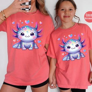 May include: Two red t-shirts with a cartoon axolotl wearing a sparkly crown. The axolotl is pink and purple with iridescent scales. The text "COLOR: WATERMELON" is in the top right corner of the image.