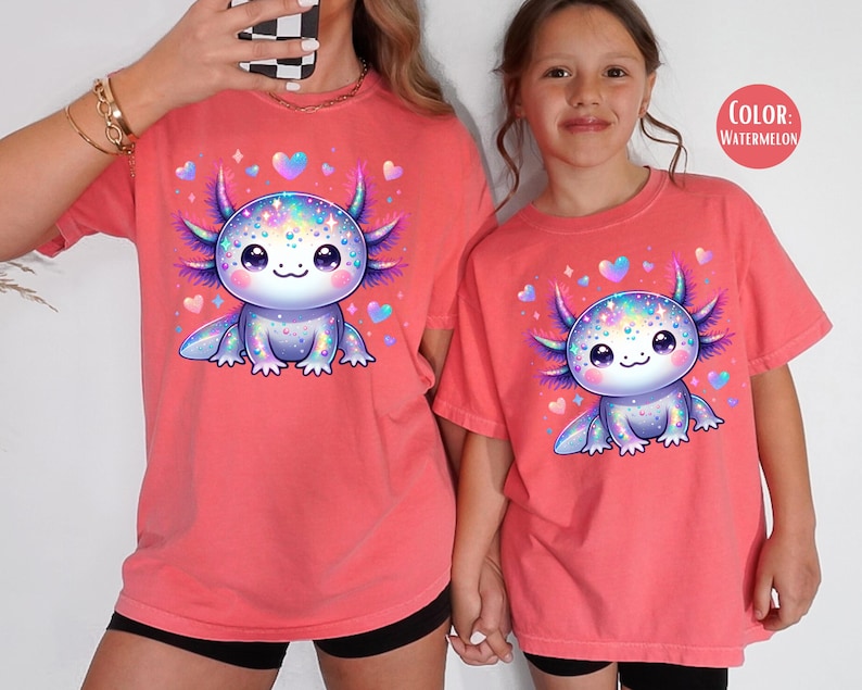 May include: Two red t-shirts with a cartoon axolotl wearing a sparkly crown. The axolotl is pink and purple with iridescent scales. The text "COLOR: WATERMELON" is in the top right corner of the image.