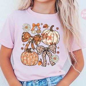 Peut inclure: T-shirt rose clair avec un motif sur le th&egrave;me de l'automne. Le design comprend des citrouilles, des n&oelig;uds, des &eacute;toiles et des &eacute;l&eacute;ments floraux dans des tons orange, marron et cr&egrave;me. Le texte "COLOR BLOSSOM" est en haut &agrave; droite.