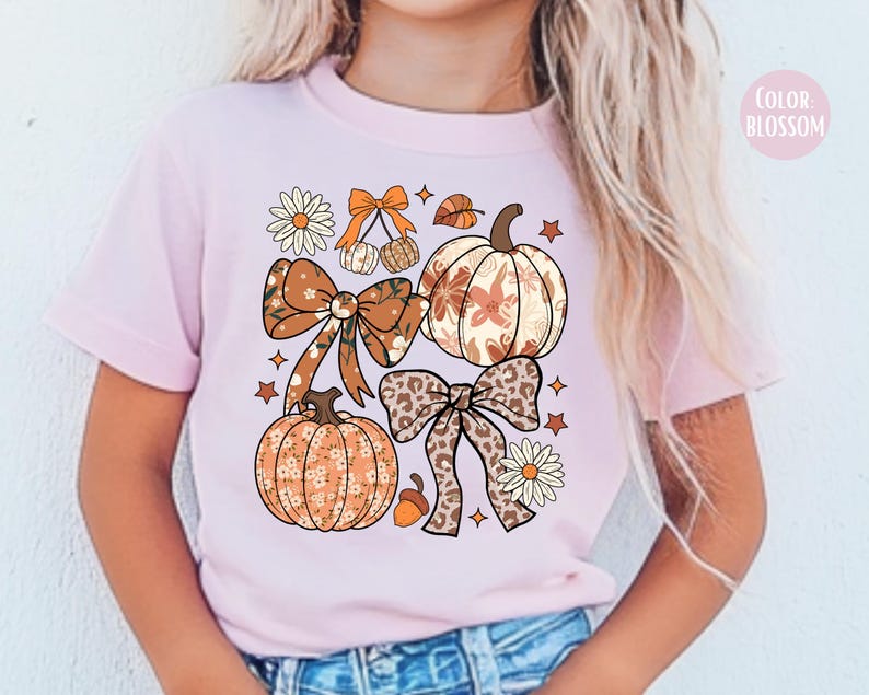 Peut inclure: T-shirt rose clair avec un motif sur le th&egrave;me de l'automne. Le design comprend des citrouilles, des n&oelig;uds, des &eacute;toiles et des &eacute;l&eacute;ments floraux dans des tons orange, marron et cr&egrave;me. Le texte "COLOR BLOSSOM" est en haut &agrave; droite.