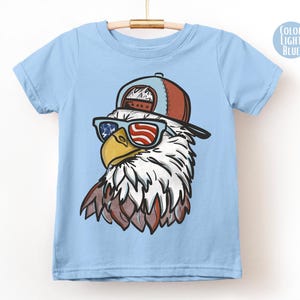 May include: Light blue t-shirt featuring a cartoon eagle wearing sunglasses with an American flag design and a brown and blue baseball cap. The eagle has white and brown feathers and a yellow beak. The shirt is hanging on a wooden hanger.