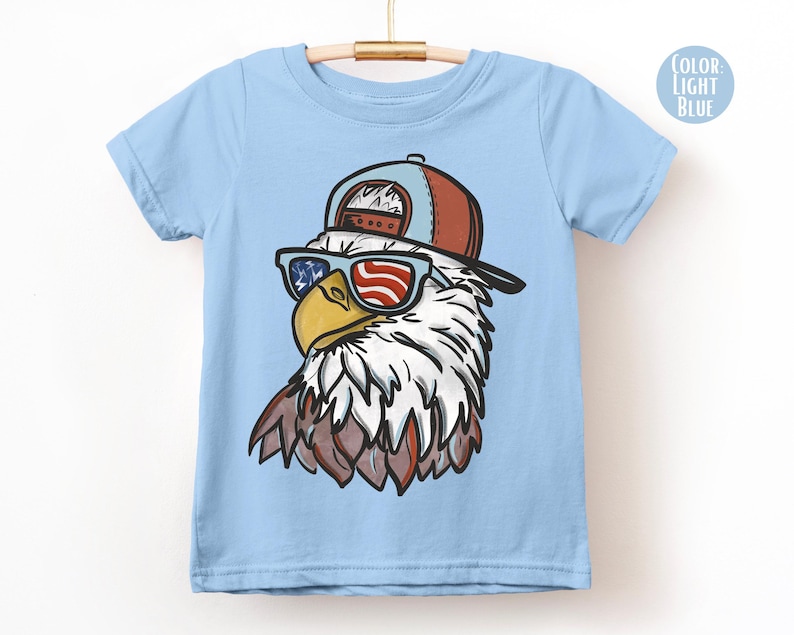 May include: Light blue t-shirt featuring a cartoon eagle wearing sunglasses with an American flag design and a brown and blue baseball cap. The eagle has white and brown feathers and a yellow beak. The shirt is hanging on a wooden hanger.