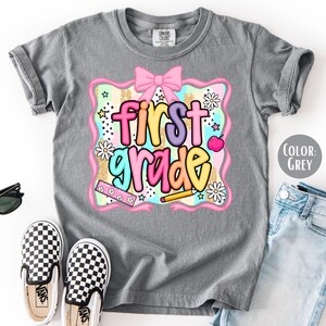 Pu&ograve; includere: Maglietta grigia con la scritta "first grade" in lettere colorate. Il design include un fiocco rosa, fiori e una matita. Sono visibili anche scarpe slip-on a scacchi bianchi e neri e occhiali da sole.