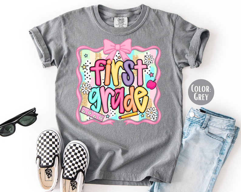 Pu&ograve; includere: Maglietta grigia con la scritta "first grade" in lettere colorate. Il design include un fiocco rosa, fiori e una matita. Sono visibili anche scarpe slip-on a scacchi bianchi e neri e occhiali da sole.