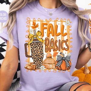 May include: Orchid-colored t-shirt with the words "FALL BASICS" in orange, along with illustrations of a leopard-print tumbler, a pumpkin, a knit hat, and fall leaves. The design is framed with a decorative border.