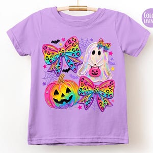 May include: A lavender short-sleeve t-shirt featuring a graphic print of a rainbow-colored pumpkin, a ghost holding a pink jack-o'-lantern bucket, and two large leopard-print bows. The design includes small bats, stars, and spider webs.