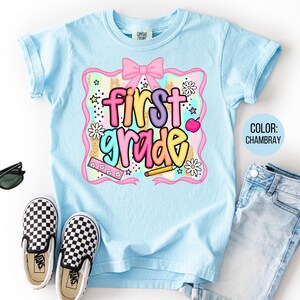 Pu&ograve; includere: Una maglietta azzurra con la scritta "first grade" in lettere colorate. La grafica include un fiocco rosa, fiori, stelle e una matita. La maglietta &egrave; abbinata a scarpe da ginnastica a scacchi bianche e nere, occhiali da sole e jeans azzurri.