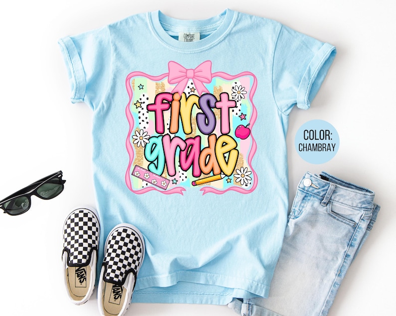 Pu&ograve; includere: Una maglietta azzurra con la scritta "first grade" in lettere colorate. La grafica include un fiocco rosa, fiori, stelle e una matita. La maglietta &egrave; abbinata a scarpe da ginnastica a scacchi bianche e nere, occhiali da sole e jeans azzurri.