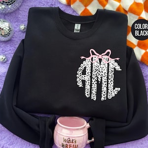 May include: Black crewneck sweatshirt with a custom monogram design in a leopard print pattern. The monogram is topped with a pink bow. The sweatshirt is displayed on a purple surface with decorative disco balls and a mug.