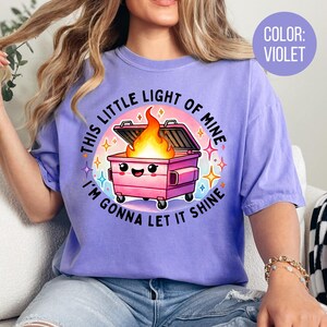 May include: A purple t-shirt featuring a graphic of a pink bin with a smiling face and flames coming out of it. The text on the shirt reads "This little light of mine, I'm gonna let it shine."