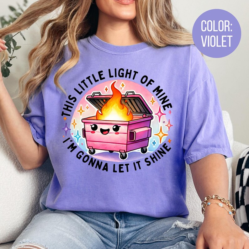 May include: A purple t-shirt featuring a graphic of a pink bin with a smiling face and flames coming out of it. The text on the shirt reads "This little light of mine, I'm gonna let it shine."