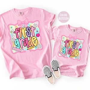 Pu&ograve; includere: Due magliette rosa con la scritta "first grade" in lettere colorate. Il design include un fiocco, stelle, fiori e una matita. Un paio di scarpe slip-on a scacchi bianchi e neri sono davanti alla maglietta pi&ugrave; piccola.