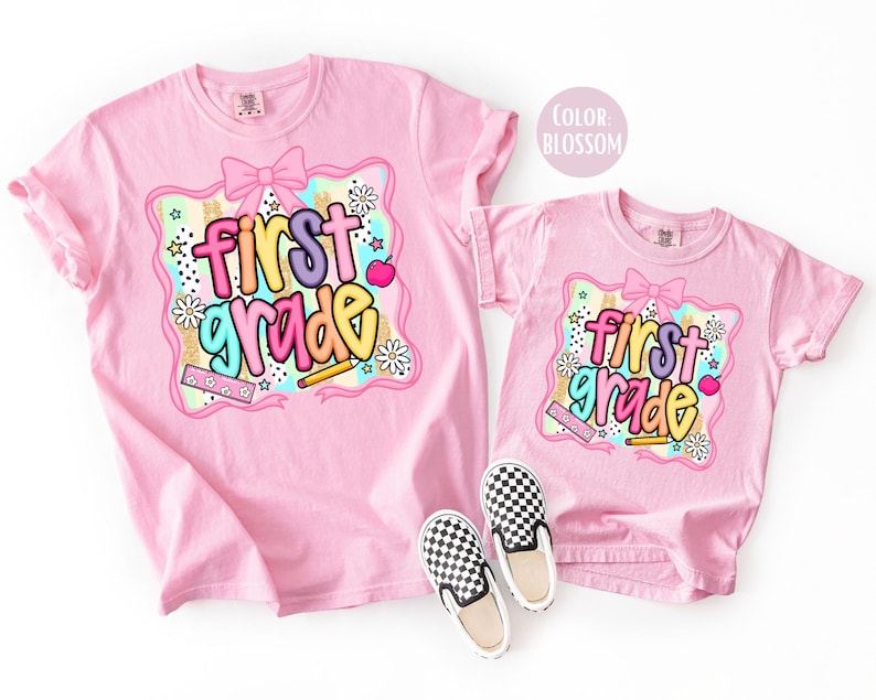 Pu&ograve; includere: Due magliette rosa con la scritta "first grade" in lettere colorate. Il design include un fiocco, stelle, fiori e una matita. Un paio di scarpe slip-on a scacchi bianchi e neri sono davanti alla maglietta pi&ugrave; piccola.
