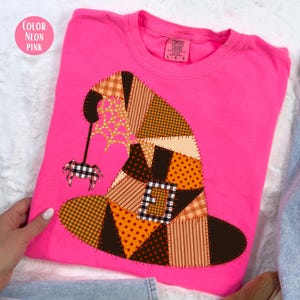 May include: A neon pink t-shirt featuring a patchwork witch hat design. The hat is composed of various fabric patterns in brown, orange, and tan hues, with a black spider and web detail. The shirt is a crew neck style.