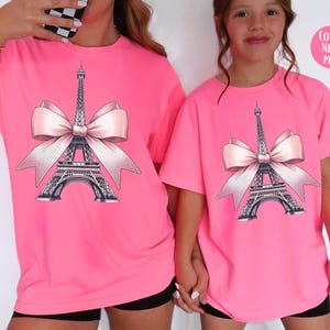 May include: Neon pink t-shirts featuring a graphic of the Eiffel Tower with a pink bow. The Eiffel Tower is detailed in black and gray. The text "COLOR: NEON PINK" is in a pink circle. The shirts are short-sleeved.