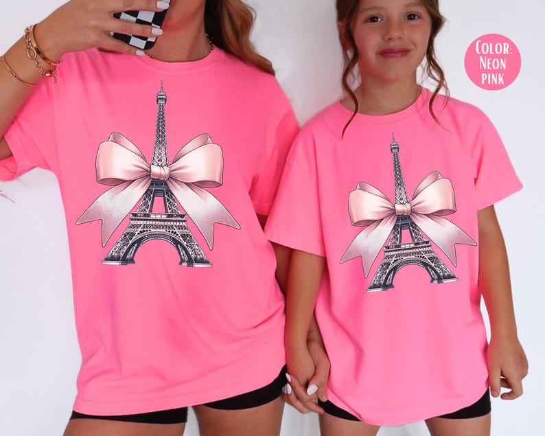 May include: Neon pink t-shirts featuring a graphic of the Eiffel Tower with a pink bow. The Eiffel Tower is detailed in black and gray. The text "COLOR: NEON PINK" is in a pink circle. The shirts are short-sleeved.