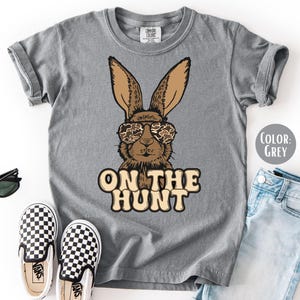 May include: A grey t-shirt with a cartoon rabbit wearing camouflage sunglasses and the text "ON THE HUNT". The image also shows black sunglasses, checkered slip-on shoes, and blue jeans.