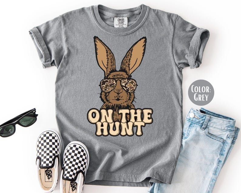 May include: A grey t-shirt with a cartoon rabbit wearing camouflage sunglasses and the text "ON THE HUNT". The image also shows black sunglasses, checkered slip-on shoes, and blue jeans.