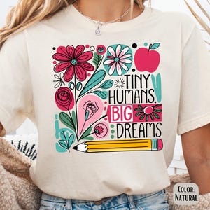 May include: A white t-shirt with a colorful graphic design featuring flowers, a pencil, and the text "Tiny Humans. Big Dreams."