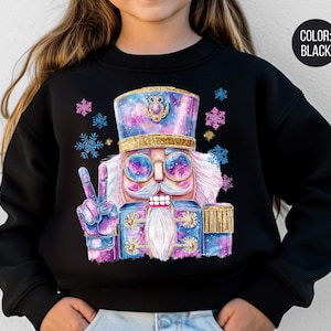 May include: Black sweatshirt featuring a colorful nutcracker design. The nutcracker is depicted with sunglasses, a white beard, and a peace sign. The design includes blue, purple, and gold accents, with snowflakes scattered around the image.