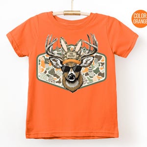 May include: Orange t-shirt featuring a deer wearing sunglasses and an "Egg Hunter" hat. The deer is set against a patterned background of Easter eggs and camouflage. The shirt is hanging on a white background.