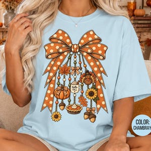 May include: A light blue t-shirt featuring a large orange polka-dot bow graphic. Hanging from the bow are various autumn-themed charms including a pumpkin, a turkey, a sunflower, a mug labeled hello fall, an acorn, and a pumpkin pie slice.