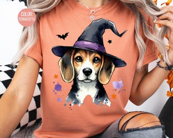 May include: Terracotta-colored t-shirt featuring a watercolor illustration of a beagle wearing a witch hat. The design includes a bat, stars, and purple watercolor splatters, perfect for Halloween.