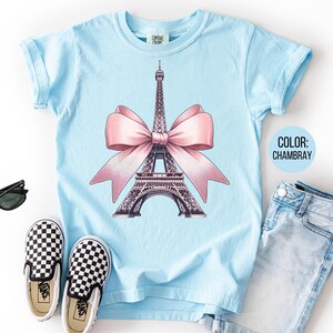 May include: A light blue t-shirt featuring a graphic of the Eiffel Tower with a pink bow. The shirt is paired with blue jeans, black and white checkered slip-on shoes, and black sunglasses. The color of the shirt is Chambray.