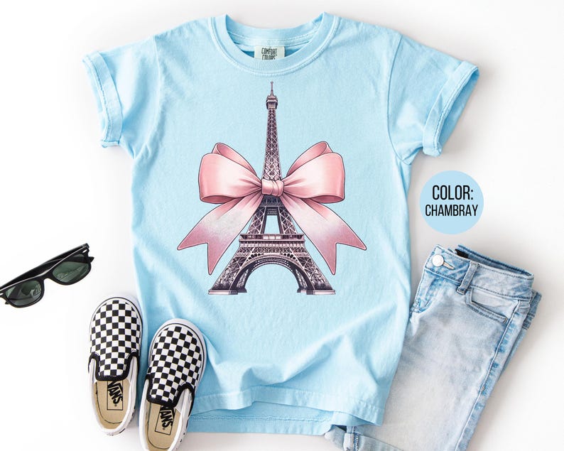 May include: A light blue t-shirt featuring a graphic of the Eiffel Tower with a pink bow. The shirt is paired with blue jeans, black and white checkered slip-on shoes, and black sunglasses. The color of the shirt is Chambray.