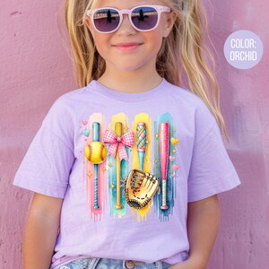 May include: A purple t-shirt with a colorful graphic of baseball bats, a ball, and a glove. The text "COLOR: ORCHID" is in the upper right corner of the image.