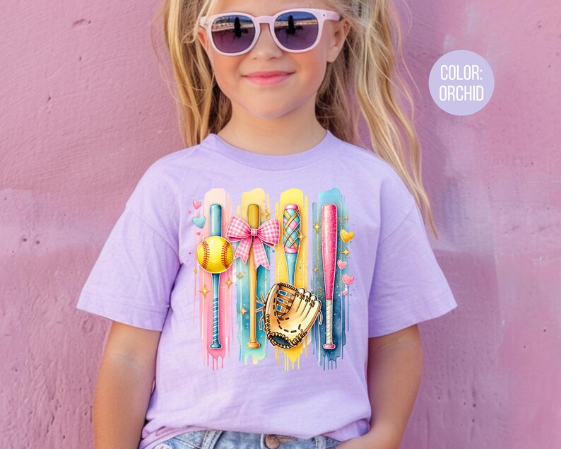 May include: A purple t-shirt with a colorful graphic of baseball bats, a ball, and a glove. The text "COLOR: ORCHID" is in the upper right corner of the image.