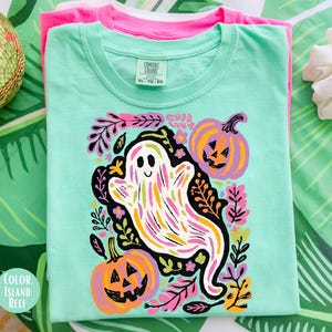 May include: A light green t-shirt featuring a colourful ghost and pumpkin design. The ghost is white with rainbow stripes, and the pumpkins are orange and purple. The shirt is folded on top of a pink t-shirt.