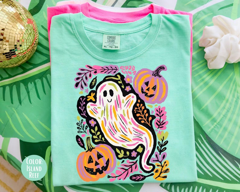 May include: A light green t-shirt featuring a colourful ghost and pumpkin design. The ghost is white with rainbow stripes, and the pumpkins are orange and purple. The shirt is folded on top of a pink t-shirt.
