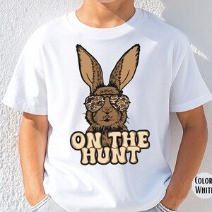May include: White t-shirt with a cartoon rabbit wearing camouflage sunglasses. The rabbit is brown with long ears. The text "ON THE HUNT" is printed below the rabbit in a tan colour.