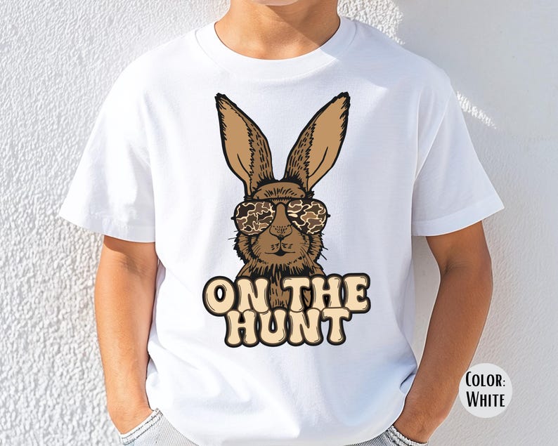 May include: White t-shirt with a cartoon rabbit wearing camouflage sunglasses. The rabbit is brown with long ears. The text "ON THE HUNT" is printed below the rabbit in a tan colour.