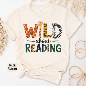 May include: A cream-colored t-shirt with the text "WILD about READING" in green and black letters. The letters are decorated with animal print patterns, including zebra, giraffe, and leopard.